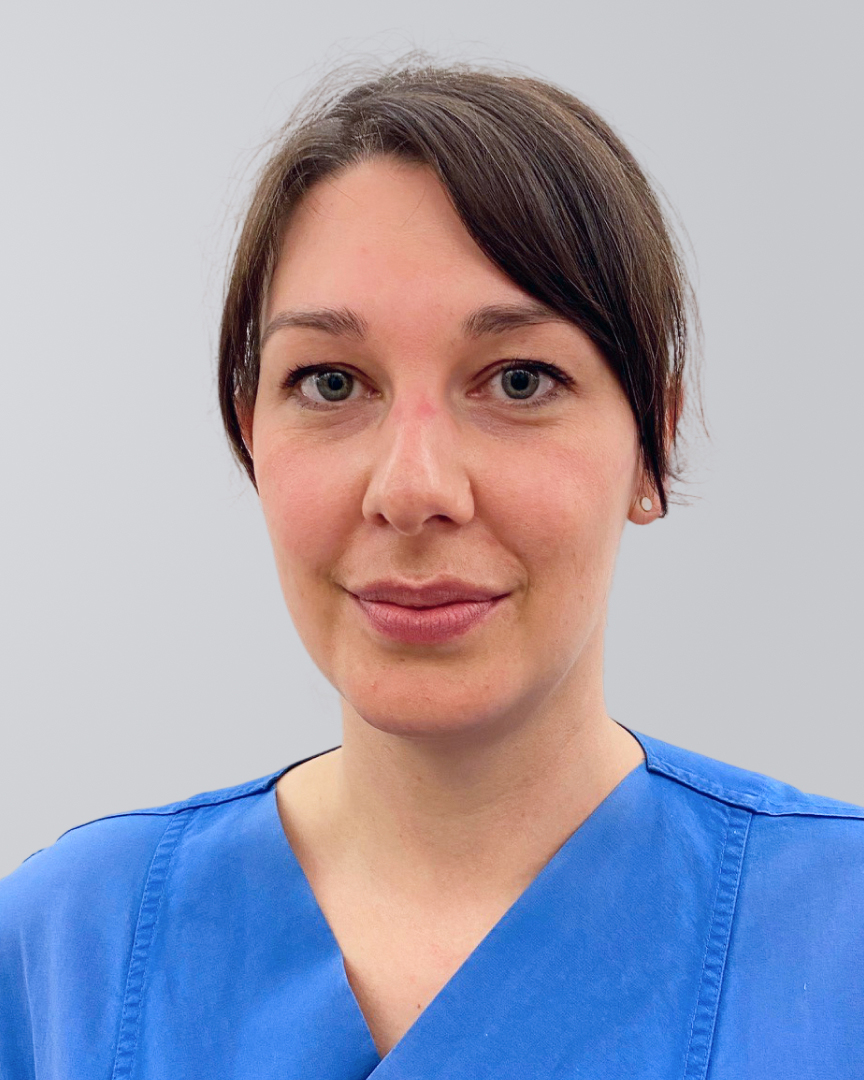 Frau mit dunklem Haar und blauer medizinischer Kleidung vor einem neutralen Hintergrund.