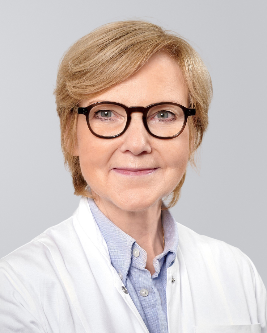 Ältere Frau mit blonden, kurzen Haaren, Brille und weißem Kittel vor neutralem Hintergrund.