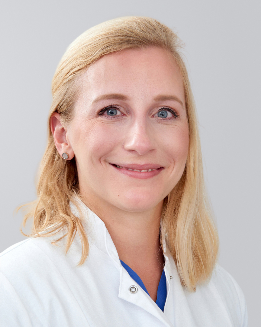 Lächelnde blonde Frau in einem weißen Kittel vor einem grauen Hintergrund.