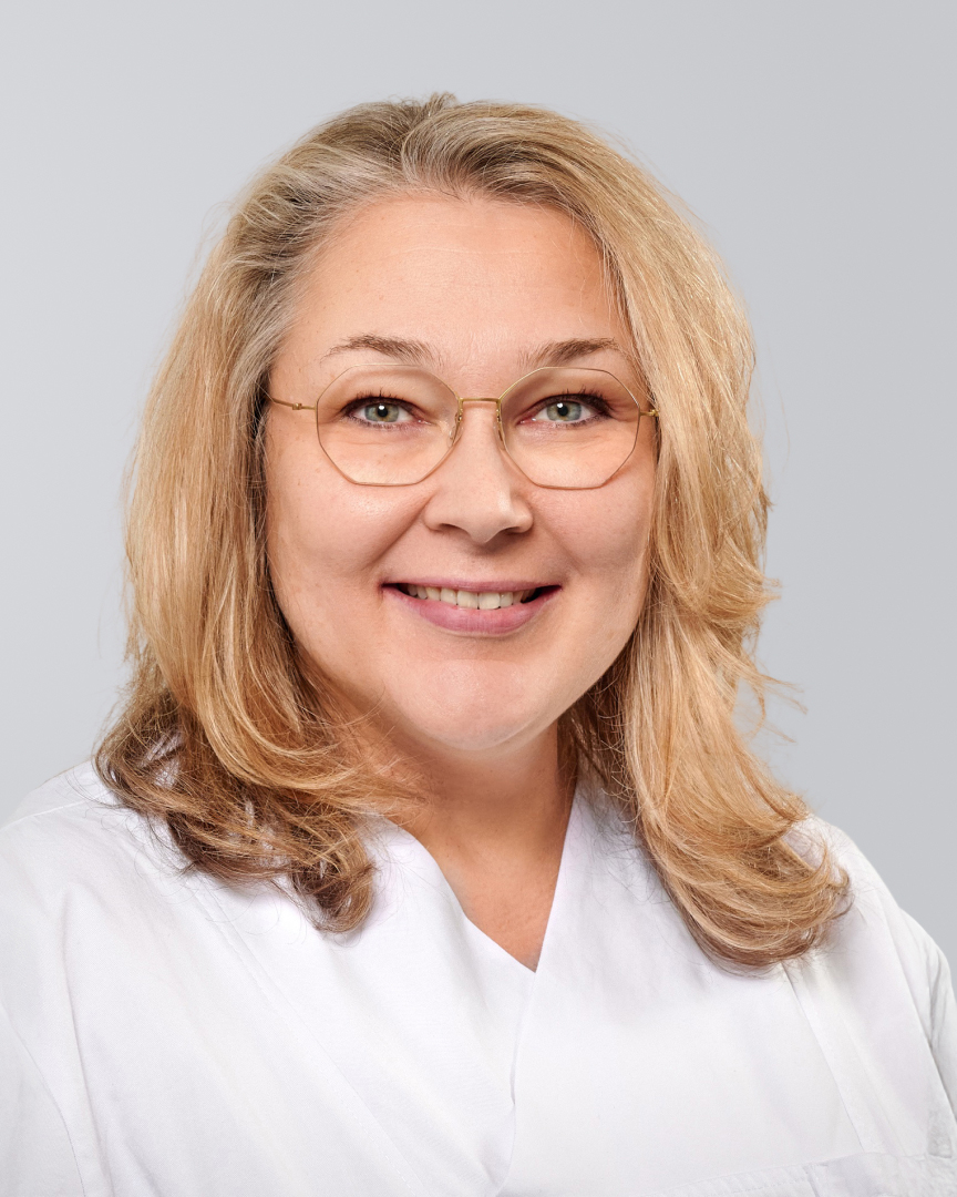 Lächelnde Frau mit blonden Haaren, Brille und einem weißen Oberteil vor einem grauen Hintergrund.