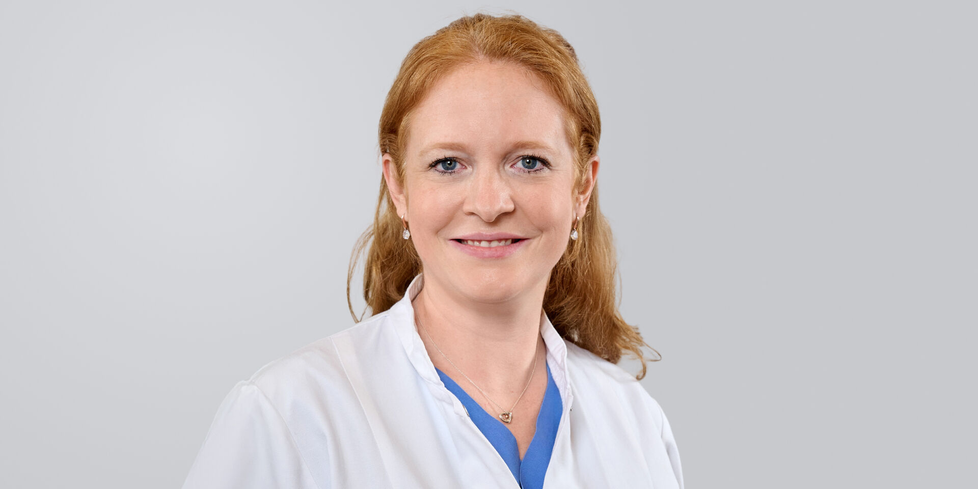 Frau mit roten Haaren in einem weißen Kittel lächelt vor einem grauen Hintergrund.