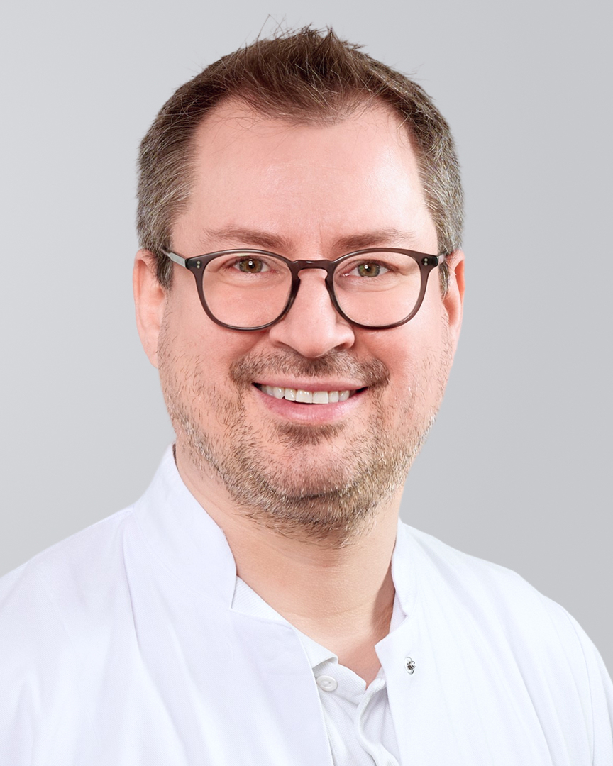 Lächelnder Mann mit Brille, Dreitagebart und weißem Kittel vor neutralem Hintergrund.