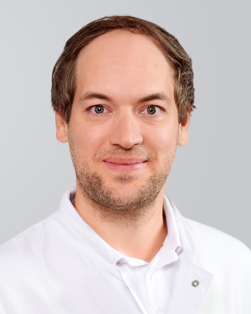 Mann mit kurzem, braunem Haar, leichtem Bart und weißem Hemd lächelt vor grauem Hintergrund.
