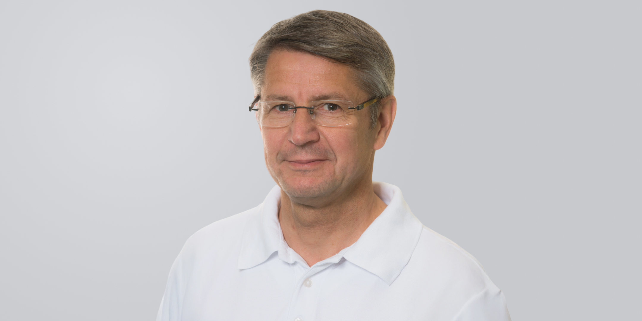 Mann mit grauen Haaren, Brille und weißem Hemd vor einem grauen Hintergrund.