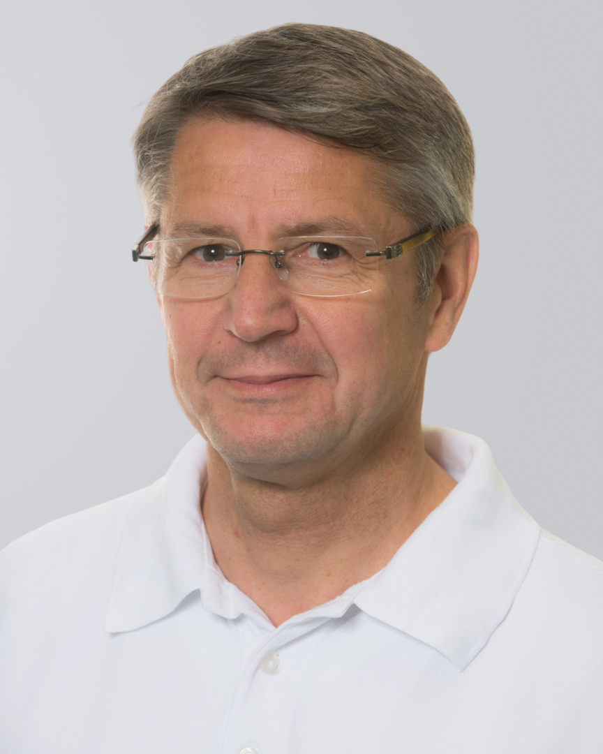 Mann mit grauen Haaren, Brille und weißem Hemd vor einem grauen Hintergrund.