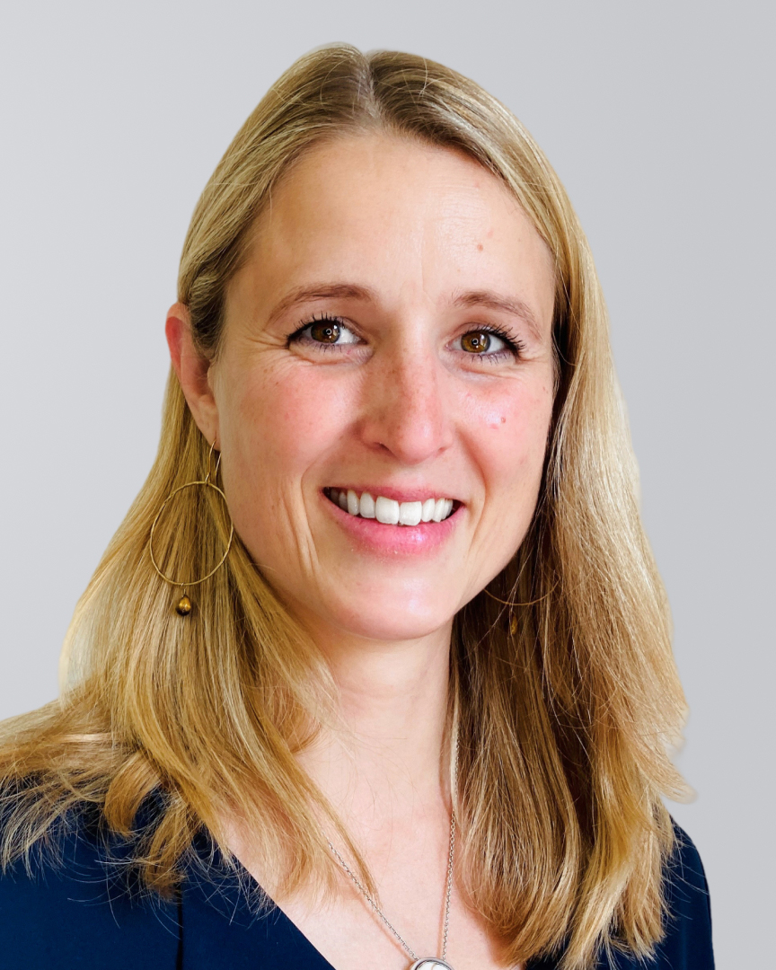 Lächelnde Frau mit langen blonden Haaren und dunkelblauem Oberteil vor einem grauen Hintergrund.