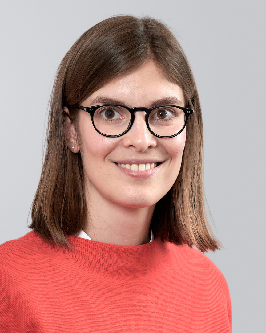 Person mit braunen, schulterlangen Haaren, rotem Pullover und Brille vor einem grauen Hintergrund.
