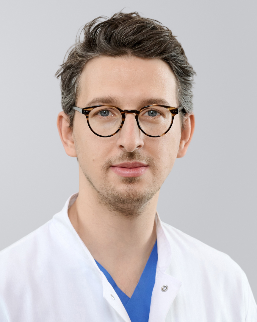 Mann in einem weißen Kittel mit Brille und blauer Kleidung vor einem grauen Hintergrund.