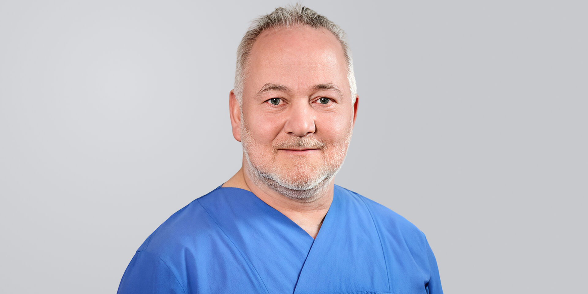 Mann mit grauen Haaren, Bart und blauer Arbeitskleidung lächelt vor einem neutralen Hintergrund.