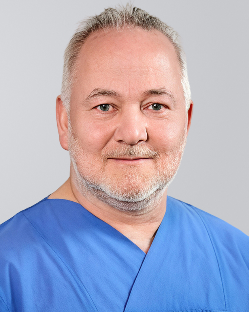 Mann mit grauen Haaren, Bart und blauer Arbeitskleidung lächelt vor einem neutralen Hintergrund.