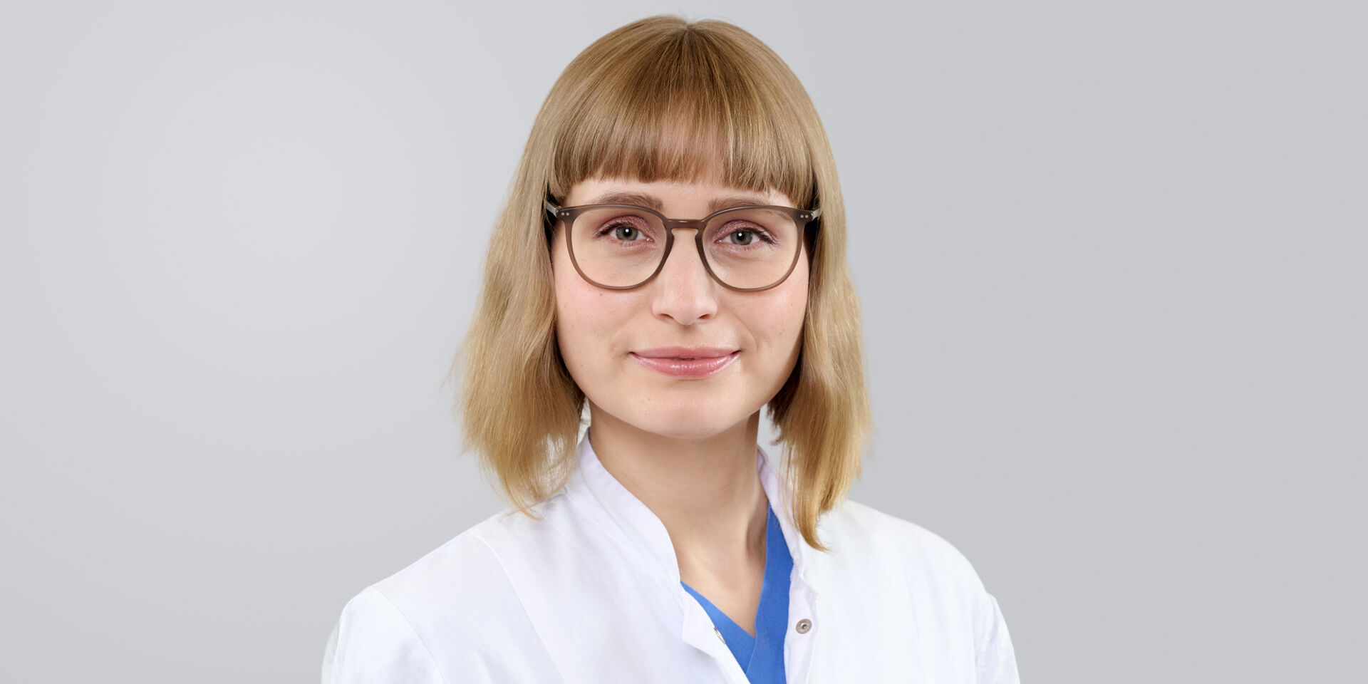 Frau mit blonden, schulterlangen Haaren, Brille und weißem Kittel vor neutralem Hintergrund.
