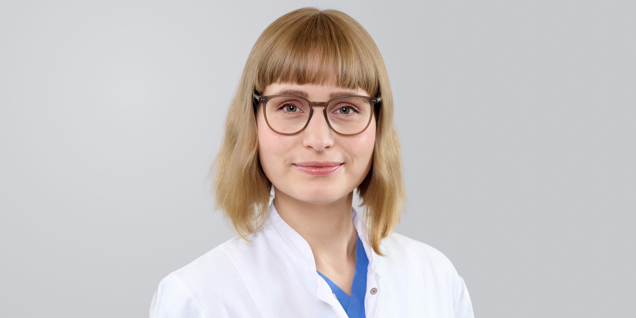 Frau mit blonden, schulterlangen Haaren, Brille und weißem Kittel vor neutralem Hintergrund.