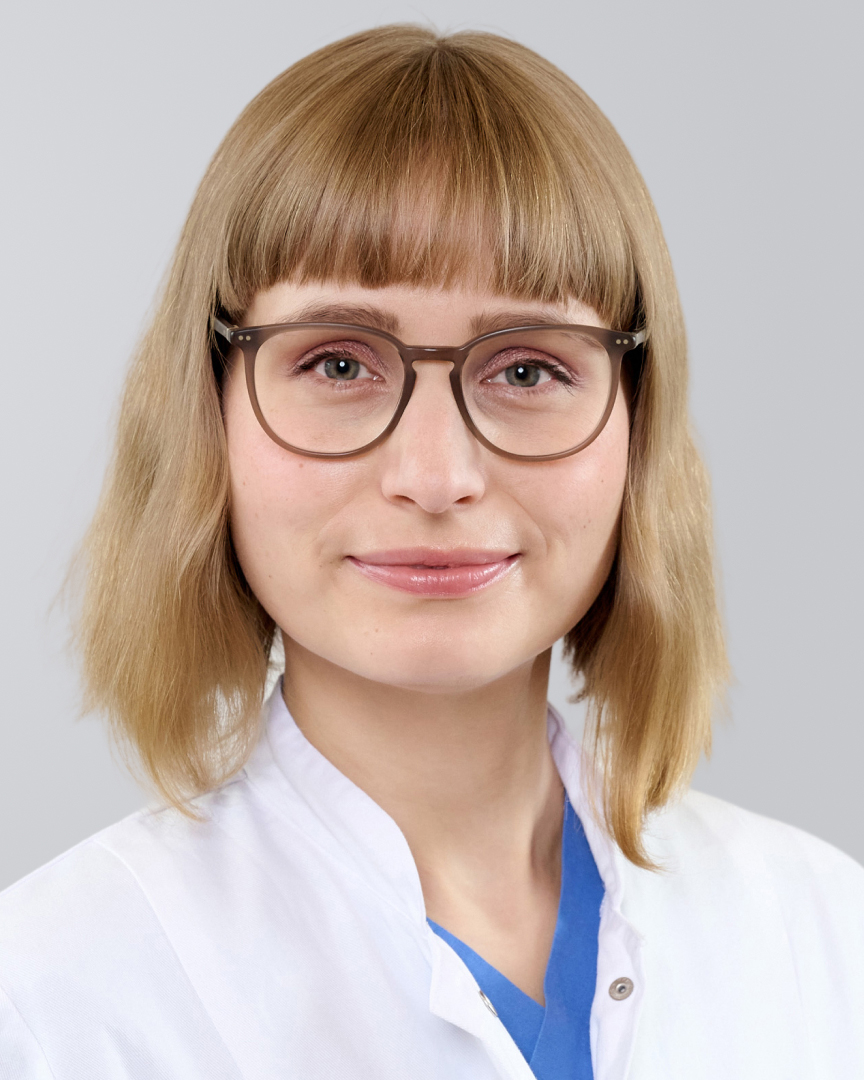 Frau mit blonden, schulterlangen Haaren, Brille und weißem Kittel vor neutralem Hintergrund.