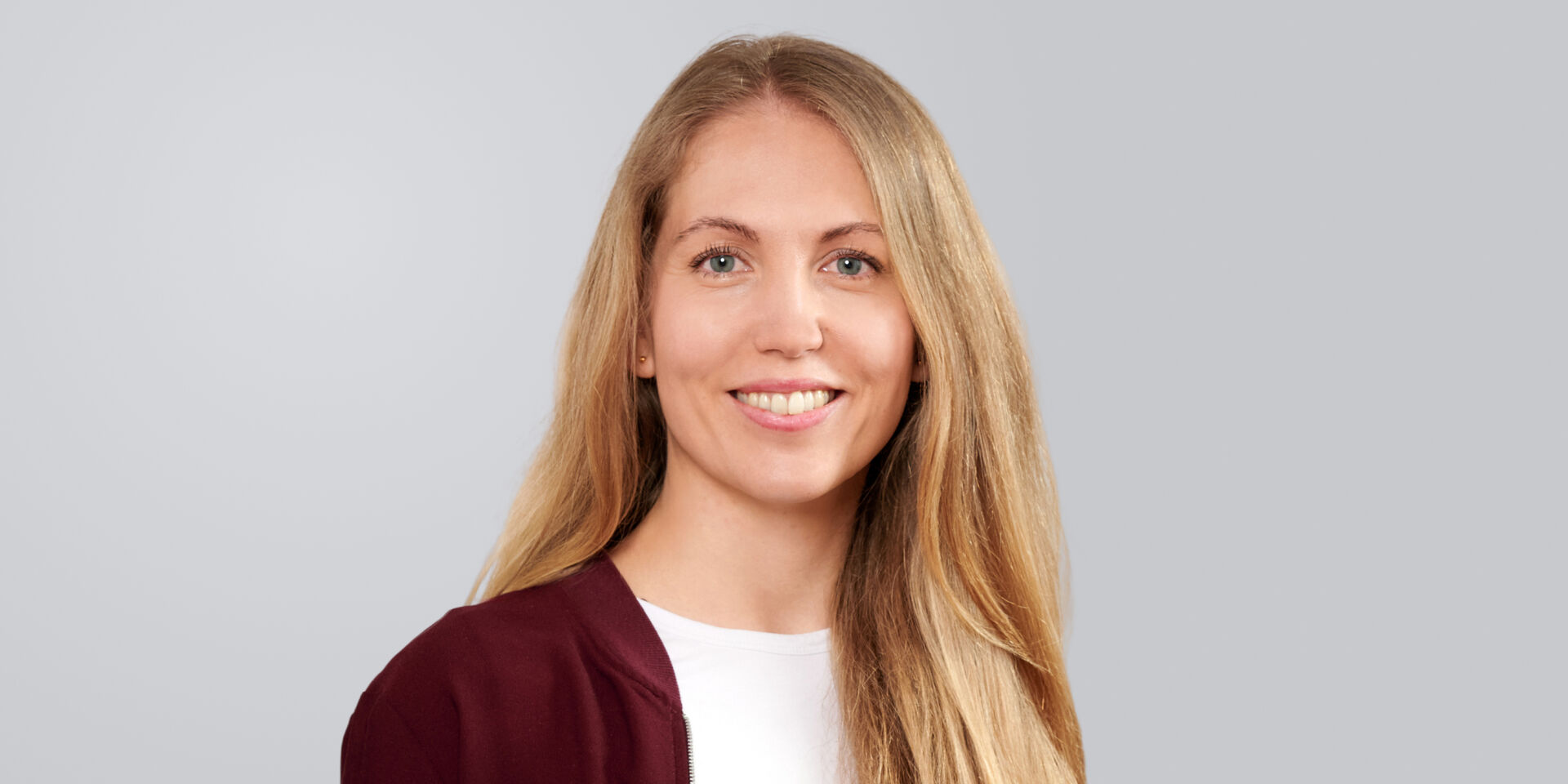 Porträt einer lächelnden Frau mit langen, blonden Haaren, die einen burgunderfarbenen Blazer trägt.