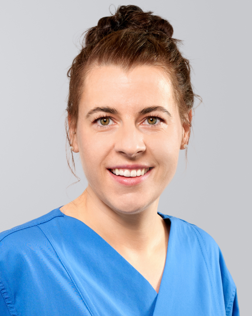 Lächelnde Frau mit dunklen, hochgesteckten Haaren und blauer Arbeitskleidung vor grauem Hintergrund.