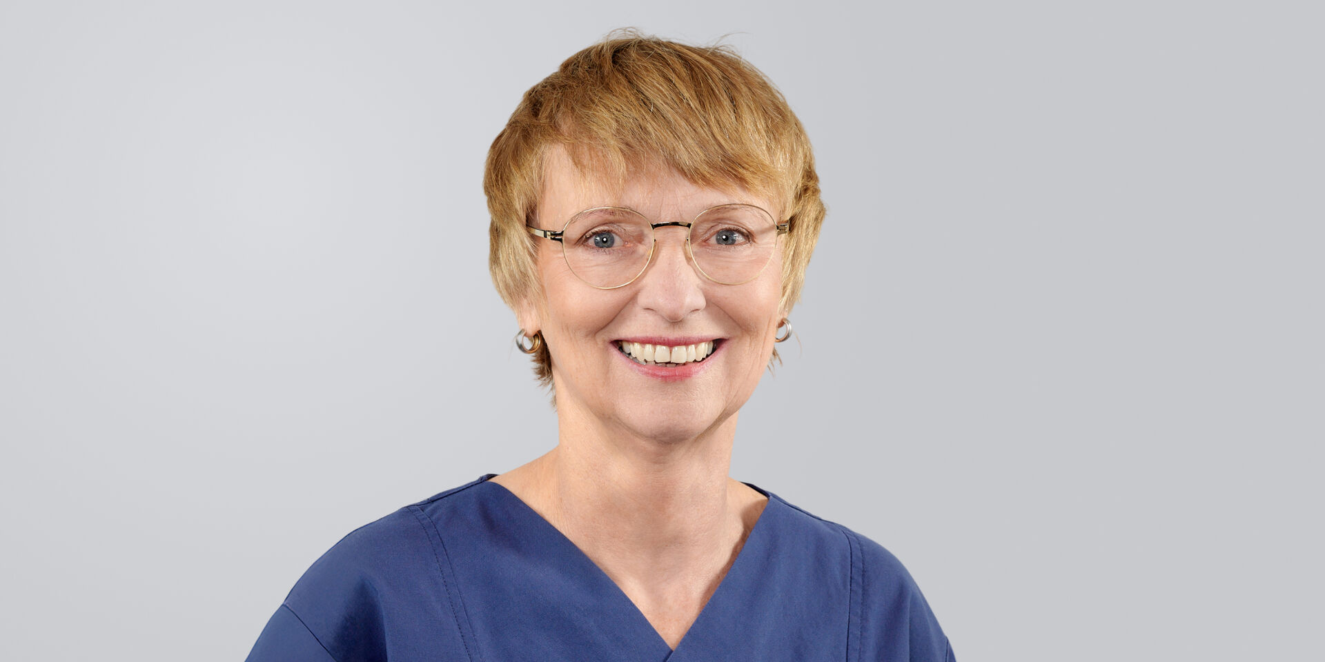 Lächelnde Frau mit kurzen, blonden Haaren, Brille und blauer Arbeitskleidung vor neutralem Hintergrund.