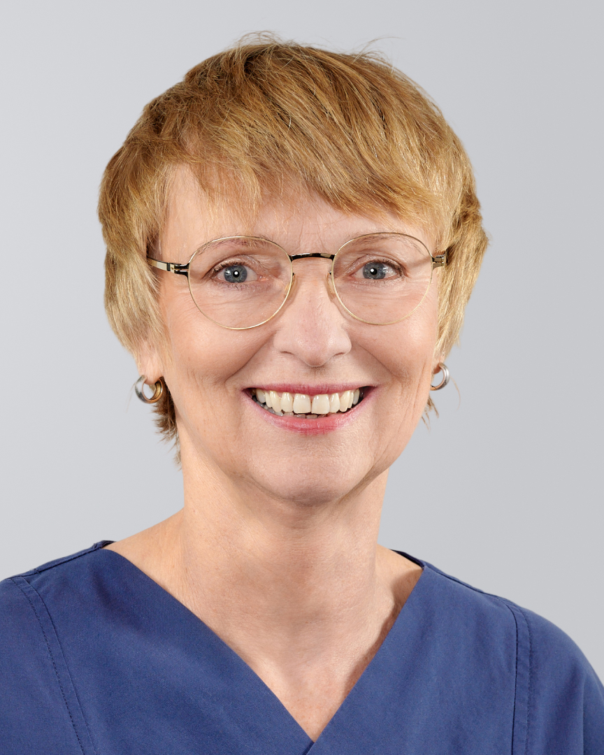Lächelnde Frau mit kurzen, blonden Haaren, Brille und blauer Arbeitskleidung vor neutralem Hintergrund.