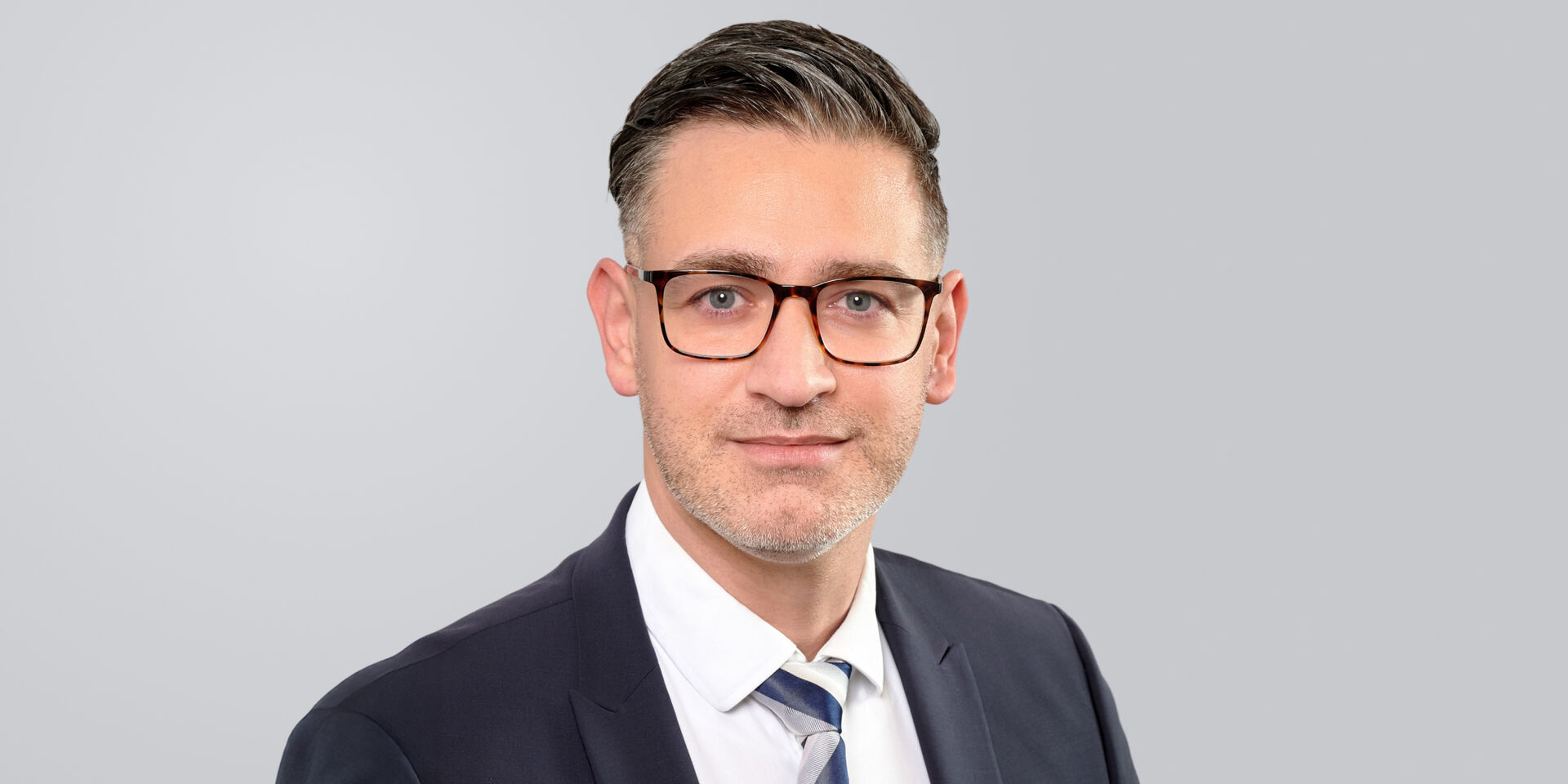 Mann mit Brille, Anzug und Krawatte lächelt vor einem grauen Hintergrund.