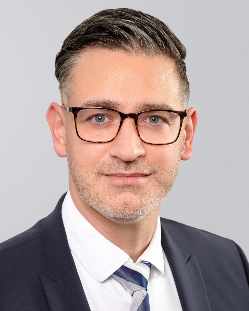 Mann mit Brille, Anzug und Krawatte lächelt vor einem grauen Hintergrund.