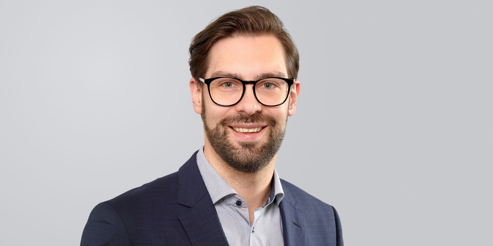 Junger Mann mit Brille, Bart und Anzug vor einem grauen Hintergrund.
