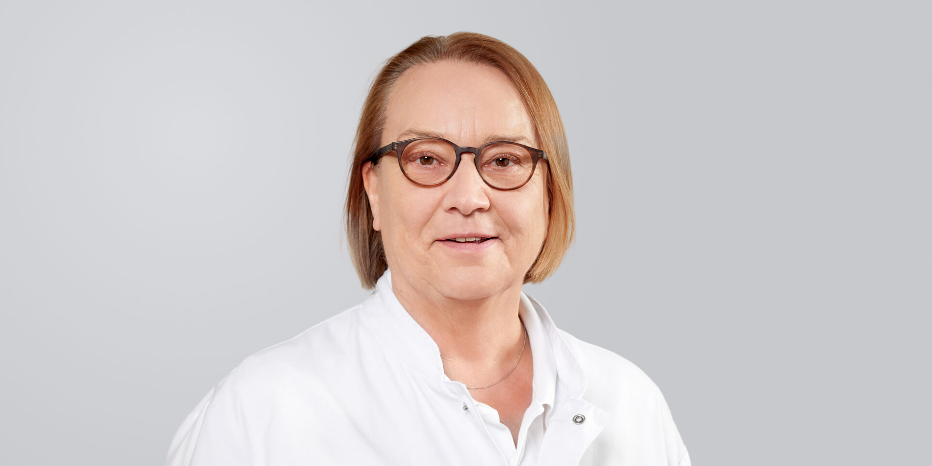 Frau mit Brille, kinnlangem, rotem Haar und weißem Kittel lächelt vor einem grauen Hintergrund.