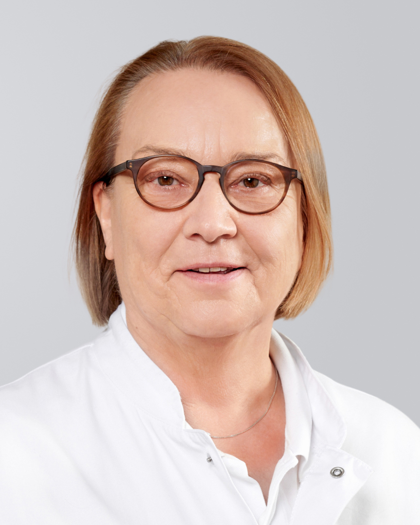 Frau mit Brille, kinnlangem, rotem Haar und weißem Kittel lächelt vor einem grauen Hintergrund.