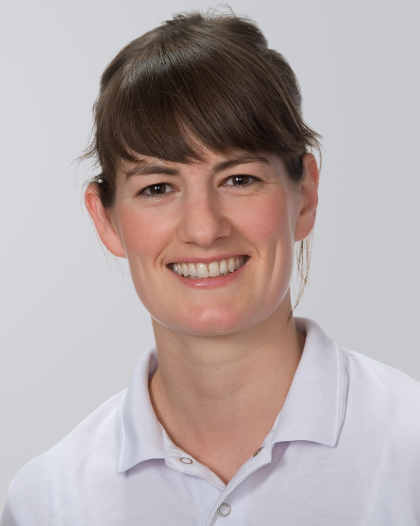 Porträt einer lächelnden Frau mit braunen Haaren und weißer Arbeitskleidung vor neutralem Hintergrund.