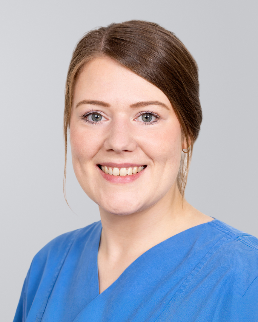 Lächelnde junge Frau in blauer Arbeitskleidung vor einem grauen Hintergrund.