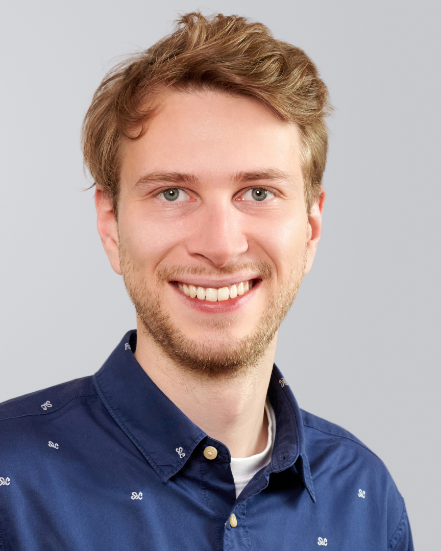 Lächelnder junger Mann mit dunkelblonden Haaren und gemustertem, blauen Hemd vor neutralem Hintergrund.