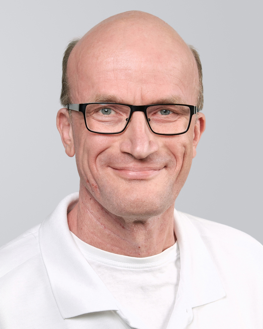 Lächelnder Mann mit Brille, Glatze und weißem Shirt vor einem grauen Hintergrund.