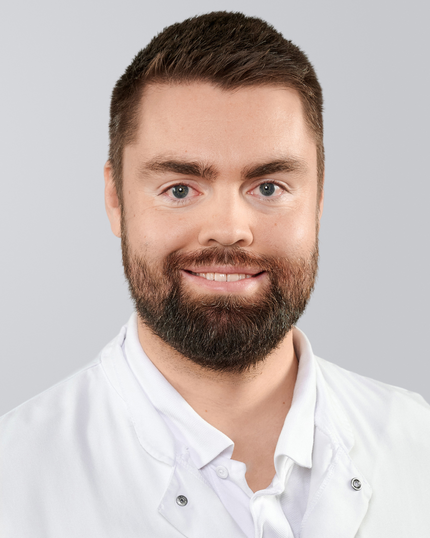Junger Mann mit Bart, braunem Haar und weißem Kittel vor grauem Hintergrund, lächelt freundlich.