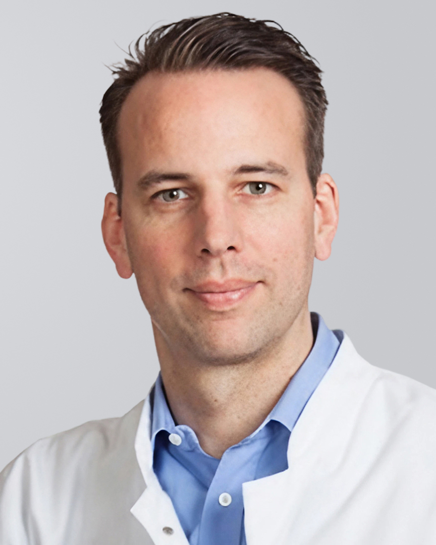 Junger Mann in weißem Kittel und blauem Hemd vor neutralem Hintergrund.