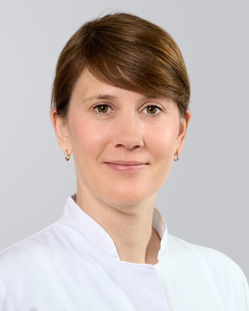 Frau mit braunen Haaren und weißem Kittel vor einem neutralen Hintergrund.