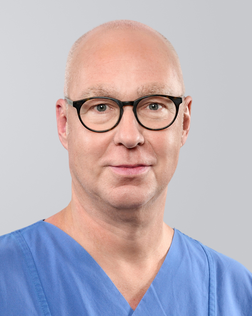 Mann mit Glatze, schwarzer Brille und blauer Arbeitskleidung Brille vor neutralem Hintergrund.