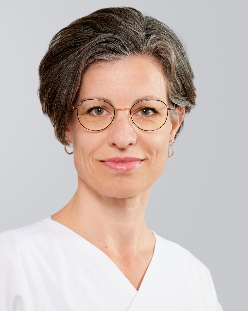 Frau mit kurzen grauen Haaren, Brille und weißem Oberteil lächelt vor einem grauen Hintergrund.