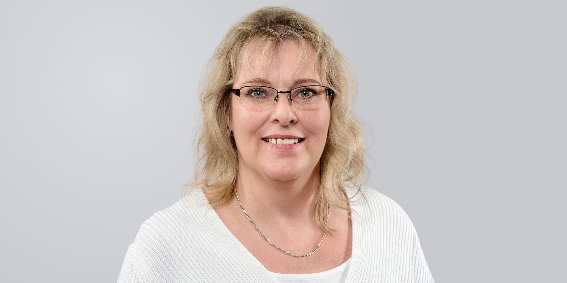 Porträt einer lächelnden Frau mit blonden, lockigen Haaren, Brille und einem weißen Pullover vor neutralem Hintergrund.