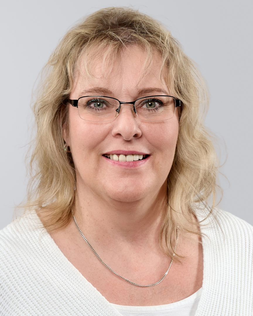 Porträt einer lächelnden Frau mit blonden, lockigen Haaren, Brille und einem weißen Pullover vor neutralem Hintergrund.
