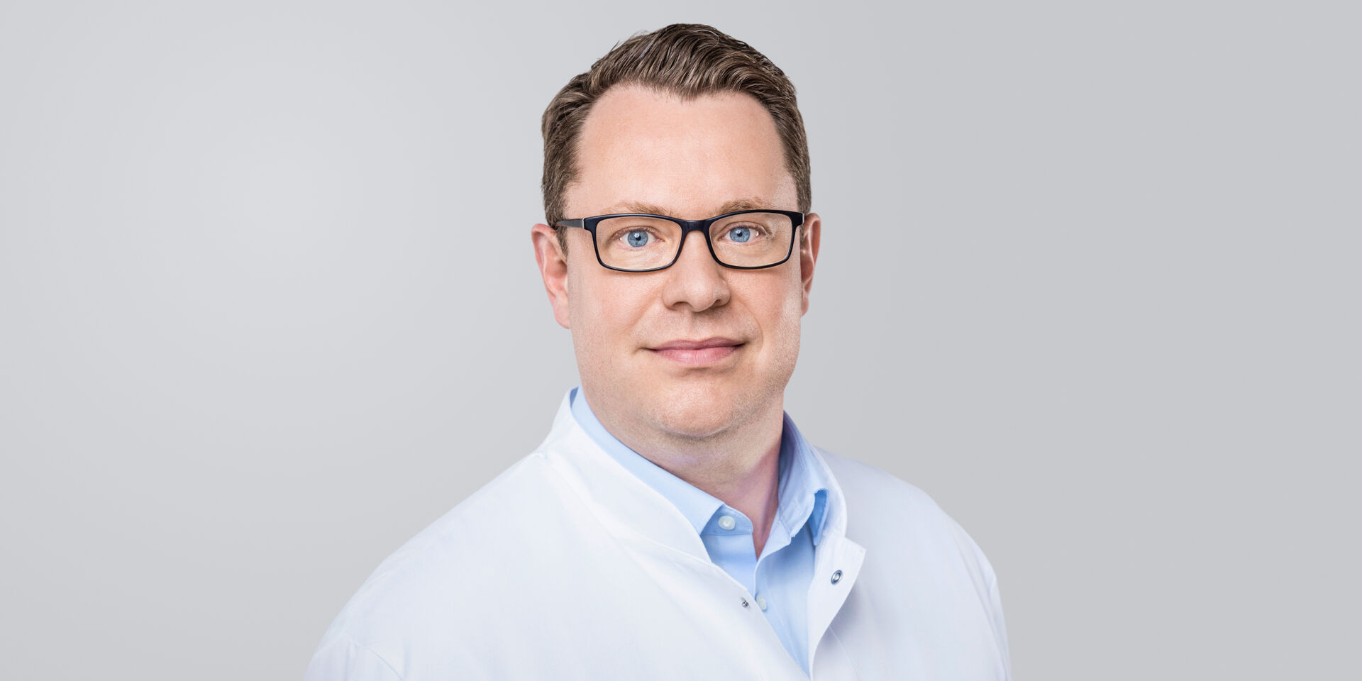 Mann mit Brille und weißem Arztkittel vor einem neutralen Hintergrund.