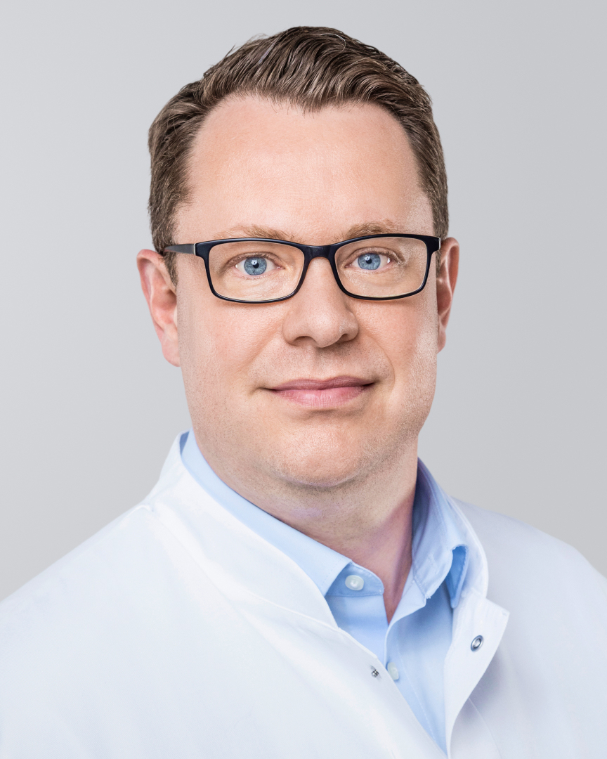 Mann mit Brille und weißem Arztkittel vor einem neutralen Hintergrund.