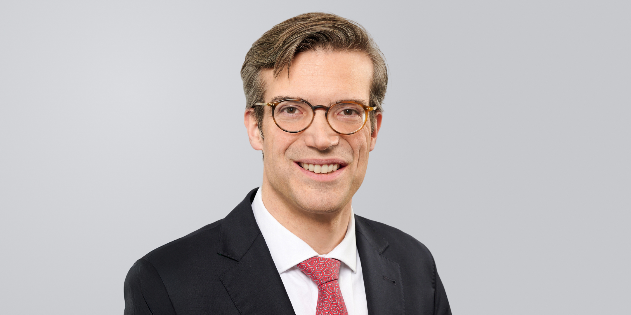 Mann mit Brille, schwarzem Anzug und roter Krawatte, lächelt vor einem grauen Hintergrund.