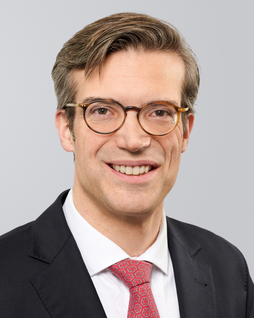 Mann mit Brille, schwarzem Anzug und roter Krawatte, lächelt vor einem grauen Hintergrund.