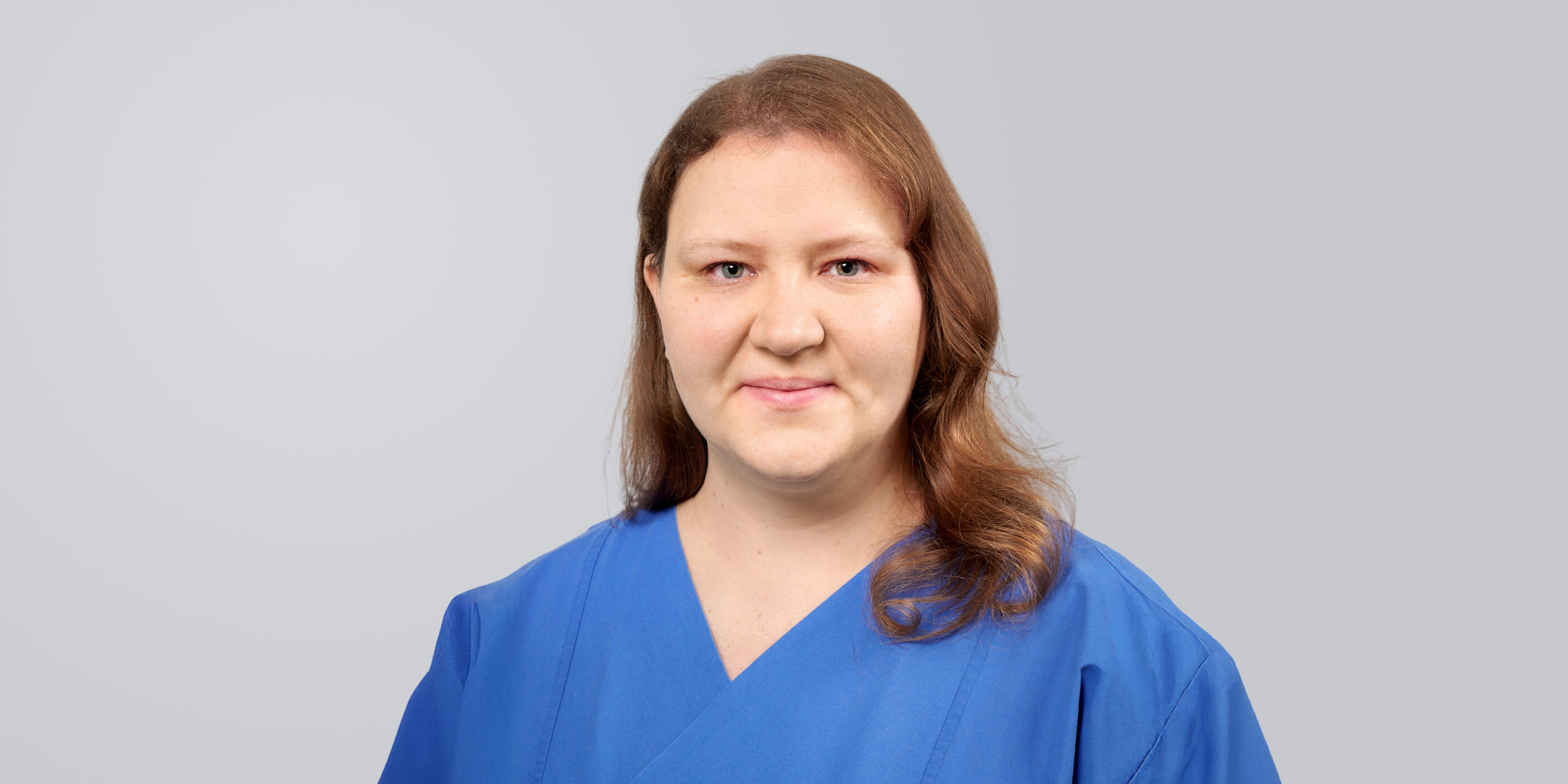 Frau mit langen, braunen Haaren in blauer Arbeitskleidung vor einem grauen Hintergrund.