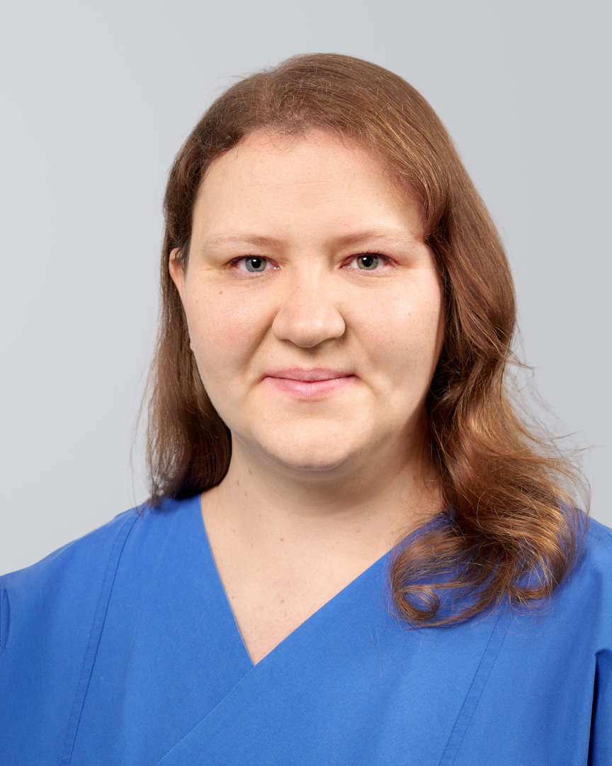 Frau mit langen, braunen Haaren in blauer Arbeitskleidung vor einem grauen Hintergrund.