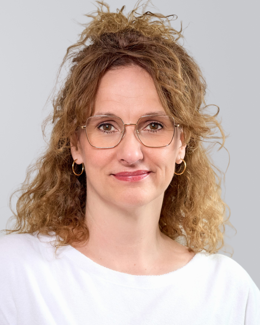 Frau mit lockigem, blonden Haar, Brille und weißem Oberteil vor einem grauen Hintergrund.