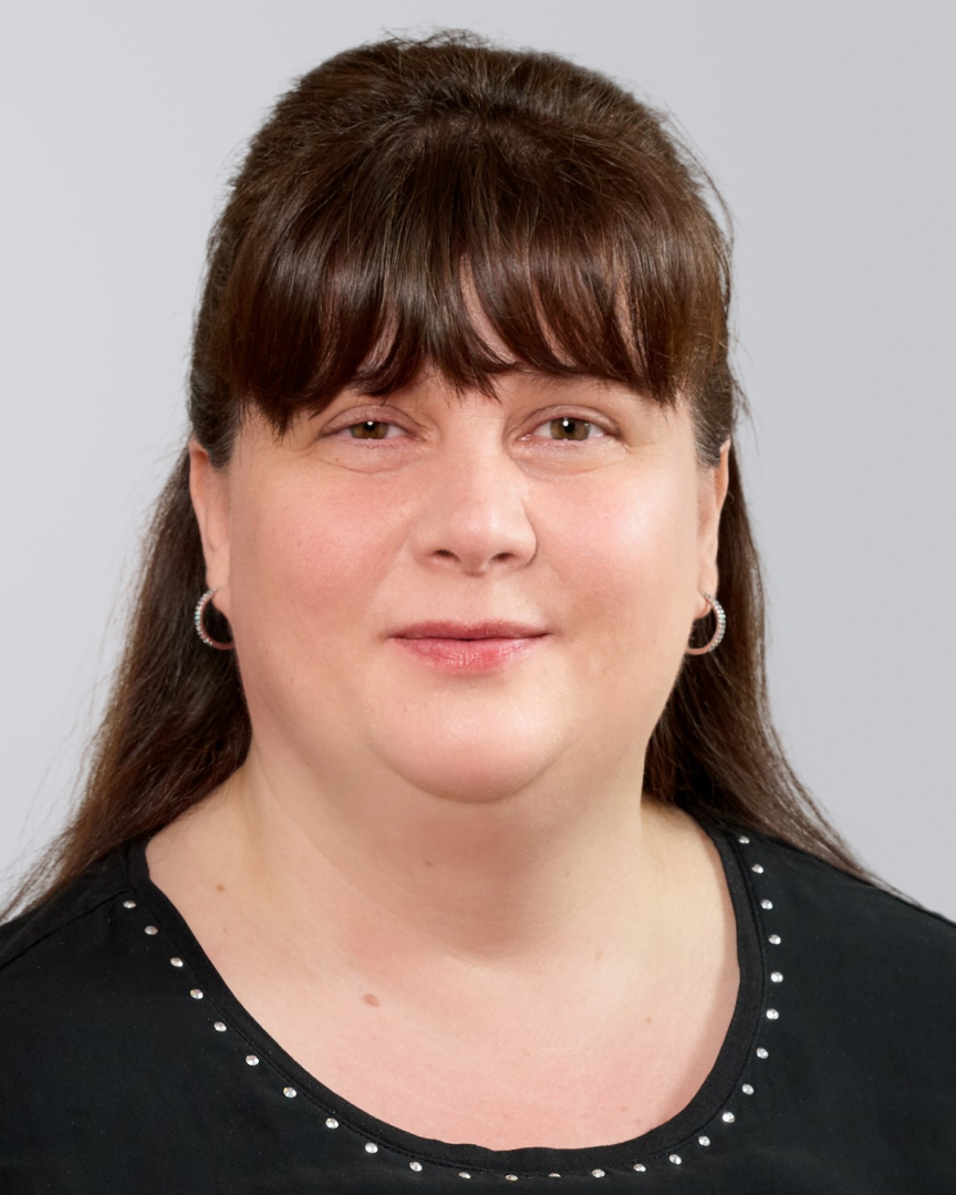Porträt einer Frau mit langen, braunen Haaren und Pony, die in einem schwarzen Oberteil vor einem grauen Hintergrund steht.