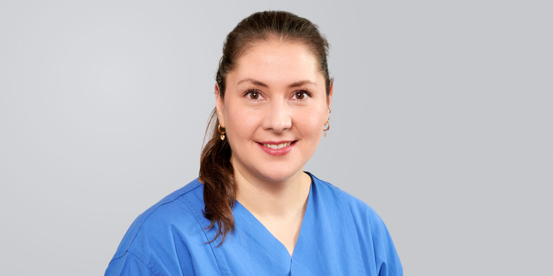 Junge Frau mit blauer Arbeitskleidung und dunklem Haar lächelt freundlich vor einem grauen Hintergrund.