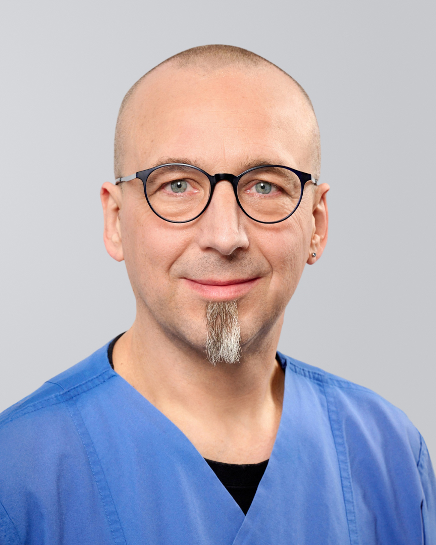 Mann mit Glatze, Brille und blauer Arztkleidung lächelt vor einem grauen Hintergrund.
