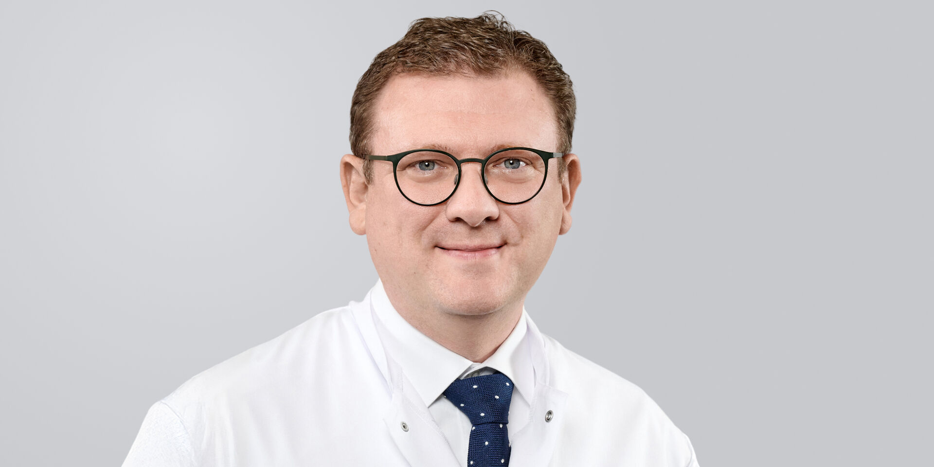 Ein lächelnder Mann mit Brille, weißem Kittel und einer blauen Krawatte vor einem grauen Hintergrund.