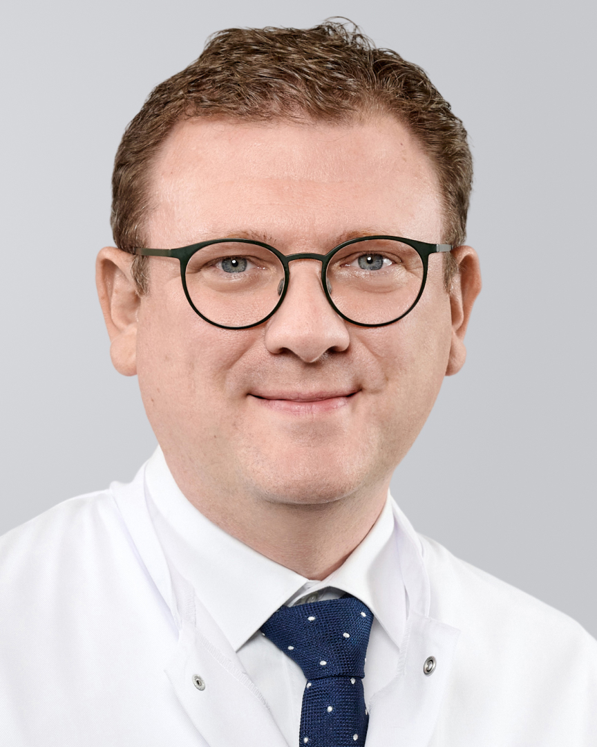 Ein lächelnder Mann mit Brille, weißem Kittel und einer blauen Krawatte vor einem grauen Hintergrund.