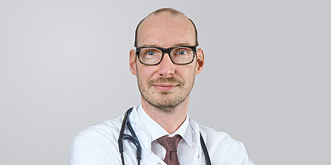 Ein Arzt mit Brille, kurzen Haaren und weißem Kittell vor einem neutralen Hintergrund.